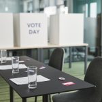Sala preparada para votação, com mesas, cadeiras e cabines de privacidade organizadas.