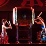 Músicos usando trajes tradicionais japoneses tocam tambores taiko durante uma apresentação em um palco.