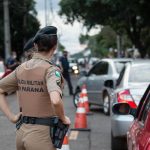Agente uniformizado controla o trânsito em uma via urbana movimentada, visto de costas.