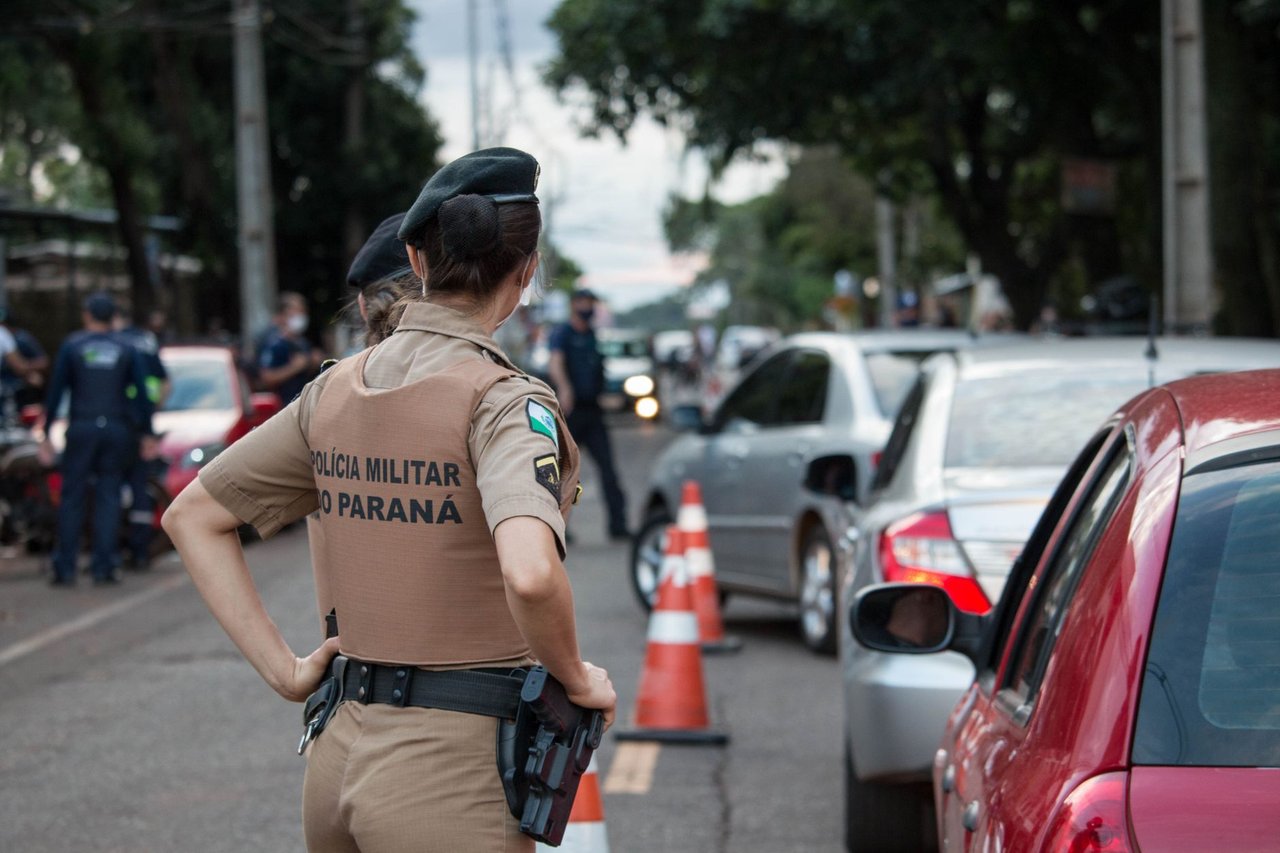 Agente uniformizado controla o trânsito em uma via urbana movimentada, visto de costas.